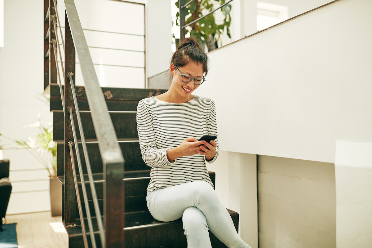 Smiling Asian businesswoman sitting on office stairs reading texts